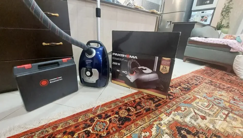 جاروبرقی پاکشوما مدل Düsseldorf C1W در کنار کارتون و جعبه نگهدارنده لوازم همراه روی زمین هستند